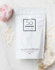 Só Luxury Coconut and Oat Milk Bath 300g pouch with scattered product around it and a pink crystal on the top left and a beige crystal on the bottom right