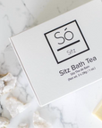 A white rectangular box of Só Sitz Bath Tea with black text, photographed on a white marble surface with a few quartz crystals nearby.
