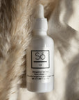 A white bottle with a dropper labeled 'Só Oil Bergamot Vanilla Face & Body Oil' styled on light coloured fabric with pampas grass.
