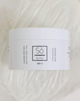 A white plastic jar of Só Butter Lavender Lime body butter, placed on top of a textured background of white whipped product
