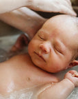 A newborn baby floating in a bath of water inside a sink, with an adult's hand gently washing the baby's hair.