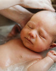 A newborn baby floating in a bath of water inside a sink, with an adult's hand gently washing the baby's hair.