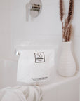 White bag with 'Só Flaky' branding on the edge of a bathtub with a cream coloured vase with dried florals.
