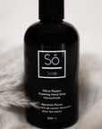 A black bottle of Só Soap hand wash concentrate in Citrus Pepper scent, standing upright styled on a blurry white background with pampas grass and cloth.
