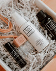 Collection of products with 'Só branding including a Lippy lip balm, small glass essential oil diffuser with a wood lid ands beige string, small peppermint hand sanitizer and a black bottle of Sunlight essential oil blend placed within a gift box with white crinkle paper.