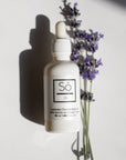 A white bottle with a dropper labeled 'Só Oil Lavender Face & Body Oil', alongside lavender flowers on a white backdrop with a shadow of the bottle.