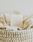 A square white bar of soap with the brand name 'Só' embossed on top, styled on a straw basket with a linen coloured cloth