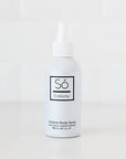 A white bottle of Só Outdoorsy Outdoor Body Spray with a white nozzle cap, standing on a white surface against a white tiled background.