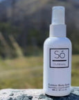 A white bottle of Só Outdoorbody spray standing on a gray stone surface with a blurred mountain landscape in the background.