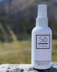 A white bottle of Só Outdoorsy outdoor body spray standing on a gray stone surface with a blurred mountain landscape in the background.