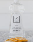 Bottle of Só Clean Citrus Rinse Aid with lemon slices on a marble surface with a blurred light background.