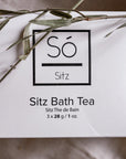 A white rectangular box of Só Sitz Bath Tea with black text, photographed on a fabric surface with natural grass elements.
