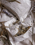 3 large tea bags styled on a linen coloured fabric one of the tea bags is sliced open showing the contents being various herbs including lavender with natural laveneder and styling elements.