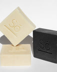 Three rectangular bars of soap branded with 'Só' on a clean white background, including one cream-colored bar standing upright, one matching cream bar lying flat, and one black bar standing upright.