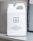 A white plastic jug of Só Clean Stain Remover & Prewash Refill bottle, displayed on a light gray shelf with a gray cylindrical container and mirror in the background.