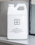 A white plastic jug of Só Clean Stain Remover & Prewash Refill bottle, displayed on a light gray shelf with a gray cylindrical container and mirror in the background.