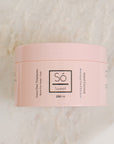 A 250 ml pink jar of Só Sweet Island Coconut body scrub, resting on a light beige marble surface.