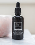 A black bottle with a dropper labeled 'Só Oil Vanilla Mint Caffeinated Face & Body Oil' with a pink quartz stone standing on a marble counter.