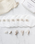 Seven white reusable cotton face pads with small sewn-on 'Só' branded tags, arranged in a row with a cream mesh bag next to a white box on a light marble surface with a few crystals.