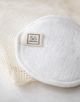 A close up of a white reusable cotton face pad with small sewn-on 'Só' branded tag on a cream mesh bag and marble surface