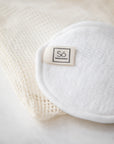 A close up of a white reusable cotton face pad with small sewn-on 'Só' branded tag on a cream mesh bag and marble surface