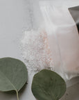 A package of Só Salty (Essential Oil) bath soak with Epsom salt and Himalayan Pink salt, displayed with a small amount of pink salt visible, surrounded by decorative greenery.