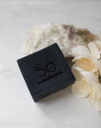 A black bar of soap branded with Só styled on a marble background with natural stone and faux florals.