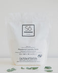 A white package of Só Clean powdered laundry soda with minimalistic design, featuring a black square logo and product information text on a light background with eucalyptus leaves.