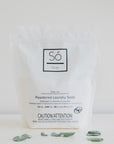 A white package of Só Clean powdered laundry soda with minimalistic design, featuring a black square logo and product information text on a light background with eucalyptus leaves.