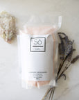 A package of Só Salty (Essential Oil) bath soak with Epsom salt and Himalayan Pink salt, scented with various essential oils and labelled Lavender Lemongrass, displayed on a light marble background with a crystal and sprigs of lavender.