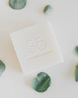 A square white bar of soap with the brand name 'Só' embossed on top, styled on a light background with scattered green eucalyptus leaves.
