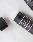 A stick of lip balm with the brand name Só Lippy placed on a surface, open, with its cap next to it, against a marble background.