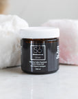 A amber glass jar with white lid of Só Silver Mission Falls Premium Ionic Silver Gel, 100 ml, set on a marble surface with white and pink rocks in the background.