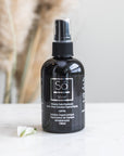 A black spray bottle of Só Silver Mission Falls Premium Ionic Silver Solution Topical Spray with a clear cap, standing on a white marble surface with a small white flower and blurred dried pampas grass in the background.