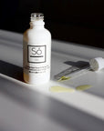 A white bottle labelled Só Oil Unscented Face & Body Oil with the dropper laying beside it containing oil, placed on a surface with a few drops of oil visible in front of the bottle.