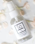A clear spray bottle of Só Magnesium Oil with a white pump and white label, photographed on a light surface with soft blurred flowers around it.