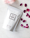 A package of Blush Sóak bath soak by Só Luxury, with a decorative pink stone and rose petals scattered around on a marble surface.