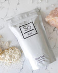 A white stand-up pouch of Só Blush Blush Soak set on a white marble surface with scattered powder, a spoonful of the powder and a decorative pink stone.