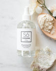 A clear spray bottle labeled 'Só Magnesium Oil' against a marble background withnatural stone and floral elements with a wooden spoon with a beige powder.