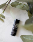 A black lip balm stick with a white logo and text, labeled 'Só Lippy Natural Lip Balm', placed on a light marble surface next to green eucalyptus leaves.