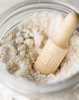 A close-up of a clear glass jar filled with a fine off-white powder, with a small light wood scoop resting inside. The scoop has an embossed 'Só' logo on it.