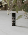 A black lip balm stick with a white logo and text, labeled 'Só Lippy Mint Lip Balm', standing on a light marble surface next to greenery.