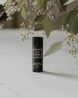 A black lip balm stick with a white logo and text, labeled 'Só Lippy Mint Lip Balm', standing on a light marble surface next to greenery.