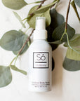 A white spray bottle labeled 'Só Outdoorsy' placed against a backdrop of green leaves, representing an outdoor body spray product.
