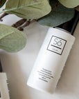 A white bottle of Só Clean peppermint hair sanitizer with a black cap, styled on a light surface with green leaves in the foreground.
