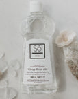 A bottle of Só Clean Citrus Rinse Aid on a marble surface styled with naturlal rocks and flowers.