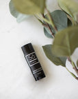 A black lip balm stick with a white logo and text, labeled 'Só Lippy Lavender Lip Balm', placed on a light marble surface next to green eucalyptus leaves.