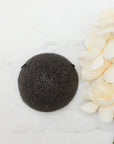 A dark charcoal gray coloured round konjac sponge arranged on a white marble surface next to cream-colored flowers.