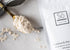 A white bag of Só Coco Oat Milk Bath styled on a marble background with a golden coloured spoon with beige powder on the spoon and black floral elements.