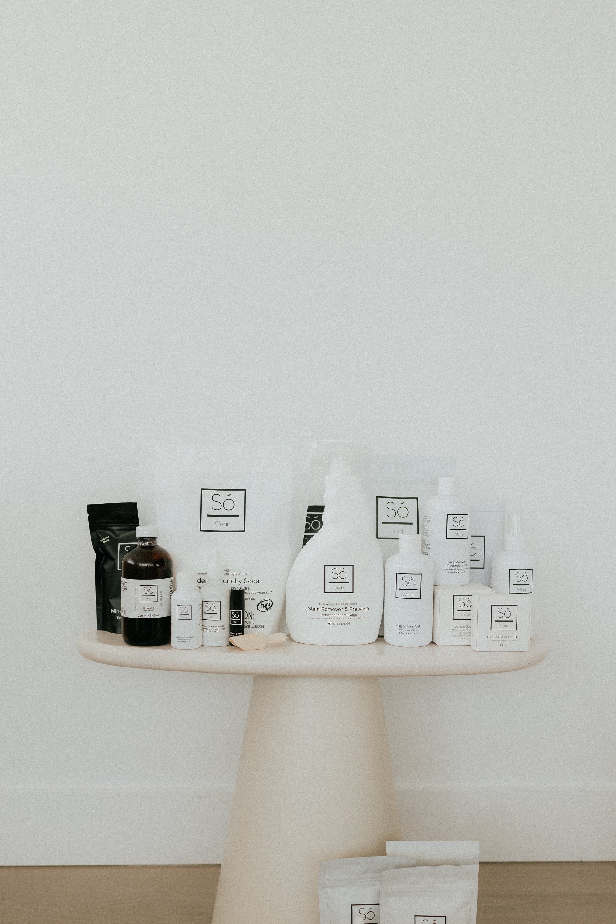 A collection of bath, body and home cleaning products from the brand Só arranged on a round, light beige pedestal table against a white wall and light wood floor.
